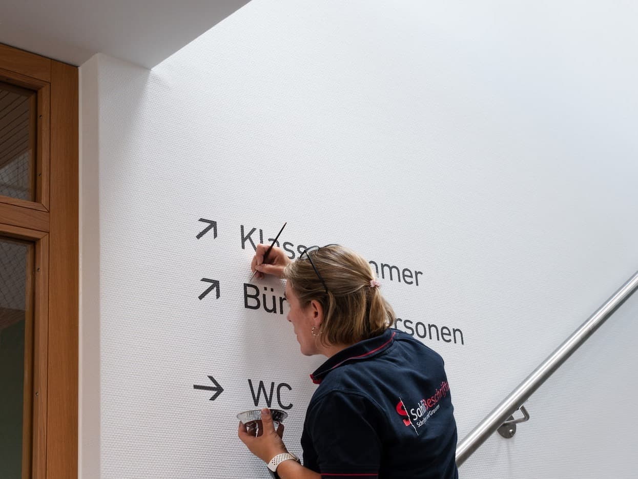 Mitarbeiterin montiert die Wegweiser-Beschriftung im Treppenhaus des Schulhauses Grüneck